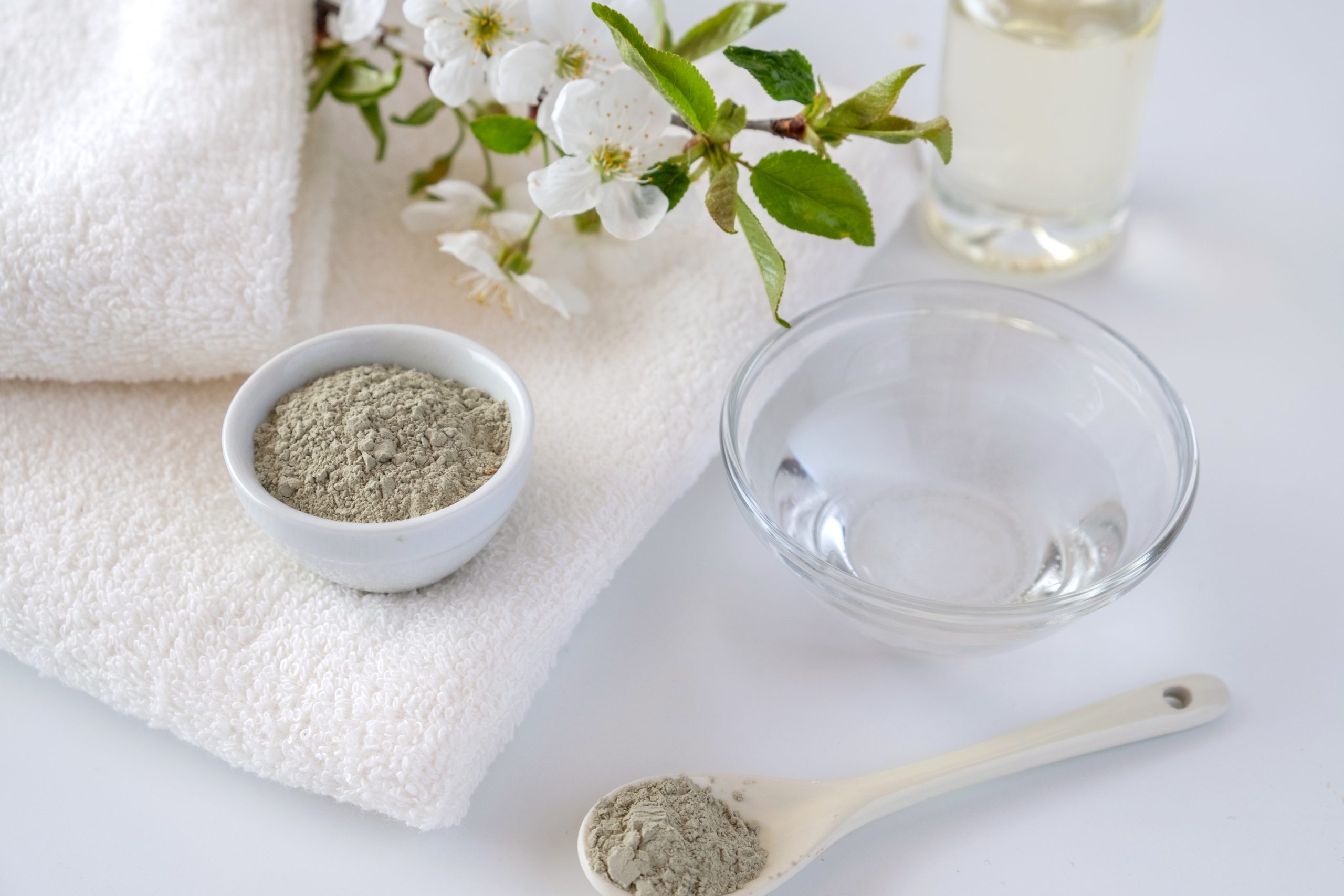 Ceramic bowl with green clay powder on white background. Ingredients for homemade facial and body mask or scrub and fresh sprig of flowering cherry. Spa and bodycare concept.
